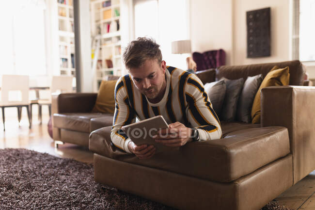 Vorderansicht eines jungen kaukasischen Mannes, der Zeit zu Hause verbringt, auf einem Sofa liegt und sein Tablet benutzt. — Stockfoto