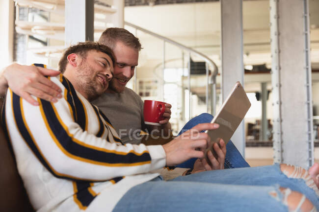 Side view of Caucasian male couple relaxing at home, sitting on a sofa, talking and smiling, holding cups of beverage while using a tablet together — Stock Photo