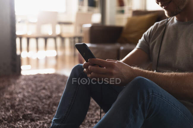 Vista laterale metà sezione di un giovane caucasico che indossa le cuffie trascorrere del tempo a casa, sdraiato su un tappeto e utilizzando il suo smartphone. — Foto stock