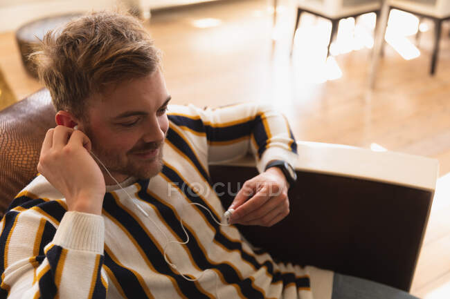 Боковой высокий угол обзора молодого кавказца надевающего наушники, проводящего время дома, сидящего на диване и улыбающегося — стоковое фото