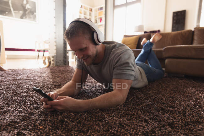 Frontansicht eines jungen kaukasischen Mannes mit Kopfhörern, der Zeit zu Hause verbringt, auf einem Teppich liegt und sein Smartphone benutzt. — Stockfoto