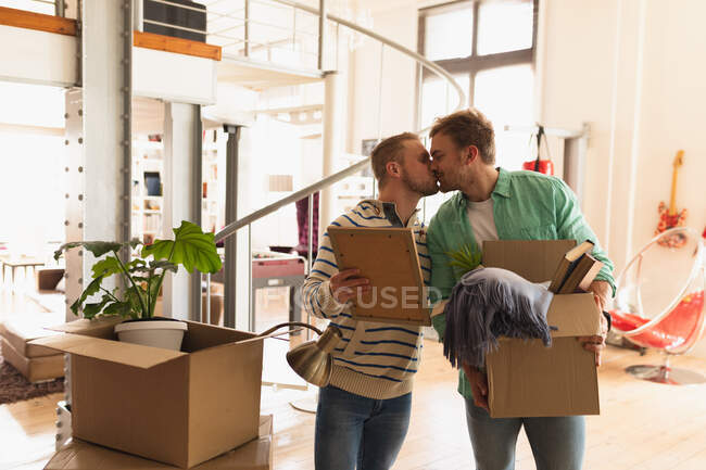 Vista frontal de una pareja de hombres caucásicos que se mudan a un nuevo apartamento, sosteniendo cajas de cartón y besando - foto de stock