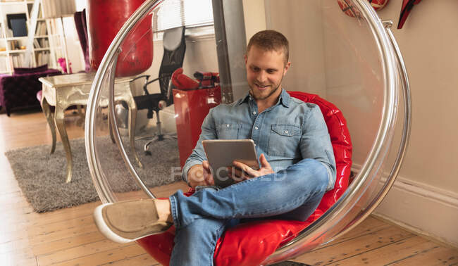 Vista frontale di un giovane caucasico che trascorre del tempo a casa, seduto su una poltrona moderna e usando il suo tablet. — Foto stock