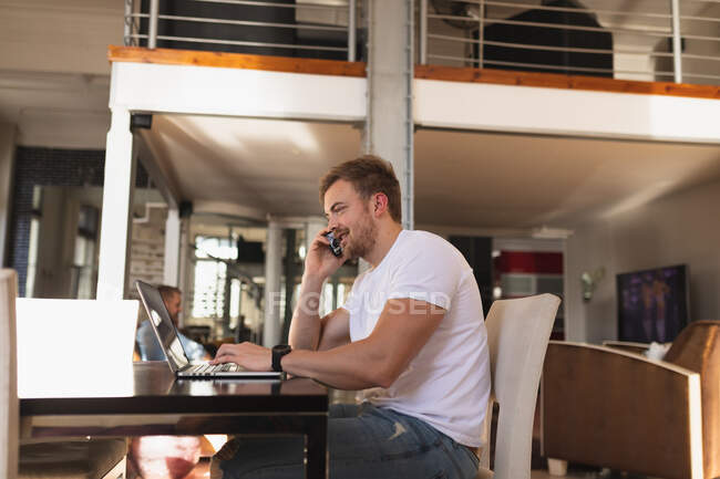 Vista lateral de un joven caucásico que pasa tiempo en casa, sentado junto al escritorio, usando su computadora portátil y hablando en su teléfono inteligente. - foto de stock