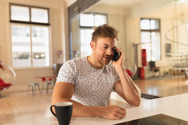 Front view of a young Caucasian man spending time at home, sitting in the kitchen with a cup of beverage, talking on the smartphone. — Stock Photo