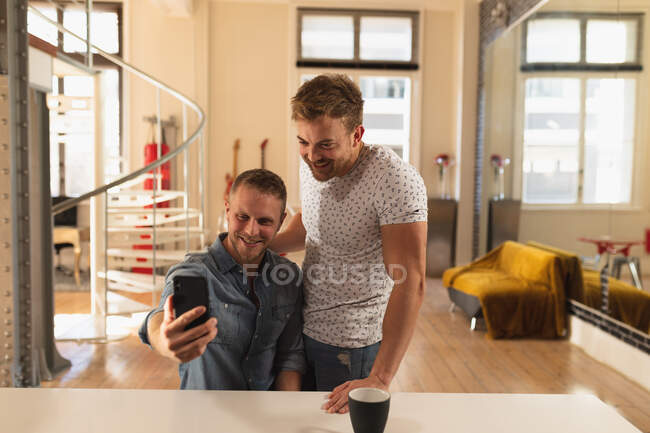 Vue de face gros plan du couple masculin caucasien se relaxant à la maison, debout dans la cuisine, embrassant, souriant et prenant selfie avec leur smartphone — Photo de stock