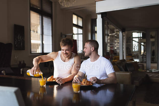 Vista frontal do casal masculino caucasiano relaxante em casa, sentado à mesa, tomando café da manhã juntos — Fotografia de Stock