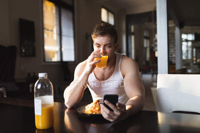 Vista frontal de un joven caucásico que se relaja en casa, sentado junto a la mesa, desayunando, bebiendo jugo de naranja y usando su smartphone - foto de stock
