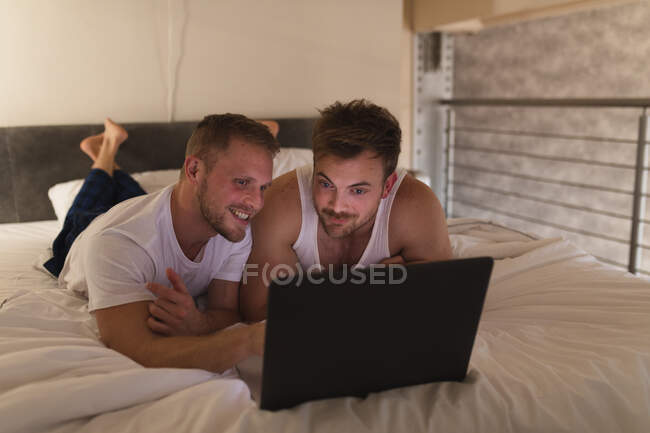 Vorderansicht eines kaukasischen männlichen Paares, das es sich zu Hause gemütlich macht, auf einem Bett liegt und mit einem Laptop interagiert — Stockfoto