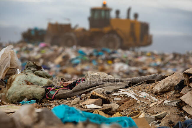 Gros plan des déchets avec bulldozer hors foyer de travail et le nettoyage des déchets empilés sur une décharge pleine de déchets avec ciel couvert nuageux en arrière-plan. Enjeu environnemental mondial de l'élimination des déchets. — Photo de stock