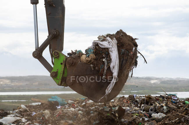 Primo piano di una scavatrice che lavora e ripulisce i rifiuti accumulati su una discarica piena di rifiuti con cielo nuvoloso coperto sullo sfondo. Questione ambientale globale dello smaltimento dei rifiuti. — Foto stock