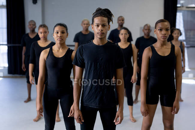 Vista frontal de un bailarín masculino moderno de raza mixta con ropa negra, parado frente a un grupo multiétnico de bailarines masculinos y femeninos en forma, mirando directamente a una cámara. - foto de stock