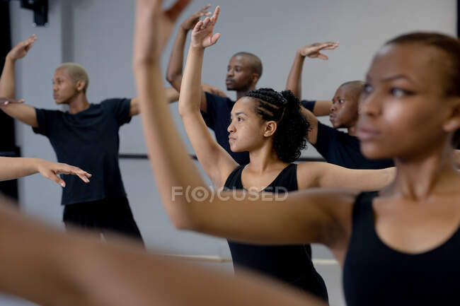 Seitliche Nahaufnahme einer multiethnischen Gruppe fitter männlicher und weiblicher moderner Tänzer in schwarzen Outfits, die während eines Tanzkurses in einem hellen Studio eine Tanzroutine praktizieren und dabei ihre rechten Arme hochhalten. — Stockfoto