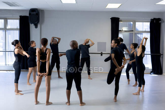 Vue arrière d'un groupe multi-ethnique de danseurs modernes, hommes et femmes, en tenue noire, pratiquant une routine de danse pendant un cours de danse dans un studio lumineux, créant un cercle et s'étirant vers le haut. — Photo de stock