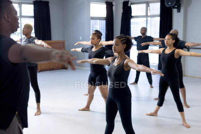 Vista frontal de un grupo multiétnico de bailarines modernos masculinos y femeninos en forma con trajes negros que practican una rutina de baile durante una clase de baile en un estudio brillante, extendiendo sus brazos. - foto de stock