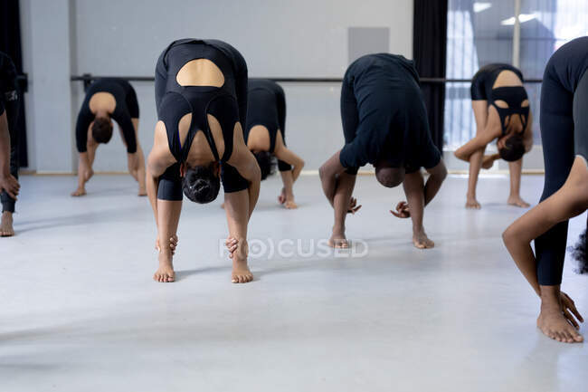 Vista frontal de um grupo multi-étnico de dançarinos modernos masculinos e femininos vestindo roupas pretas praticando uma rotina de dança durante uma aula de dança em um estúdio brilhante, esticando-se e fazendo arcos profundos.. — Fotografia de Stock