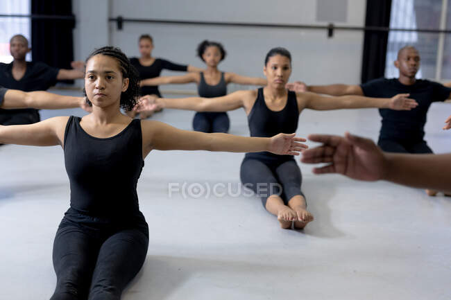 Vue latérale d'un groupe multi-ethnique de danseurs modernes en forme, hommes et femmes, portant des tenues noires pratiquant une routine de danse lors d'un cours de danse dans un studio lumineux, assis sur le sol et étirant les bras. — Photo de stock