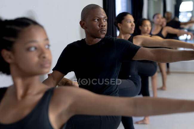 Vue de côté près d'un groupe multi-ethnique de danseurs modernes hommes et femmes en forme portant des tenues noires pratiquant une routine de danse lors d'un cours de danse dans un studio lumineux, debout près d'une main courante et s'étirant vers le haut. — Photo de stock