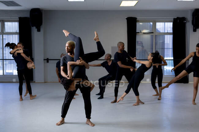 Вид спереду змішаної гонки підходять чоловічі та жіночі сучасні танцюристи в чорних костюмах, які практикують танцювальну рутину під час танцювального класу у яскравій студії, чоловік тримає жінку позуючи догори ногами, тоді як інші танцюристи стоять на задньому плані . — стокове фото