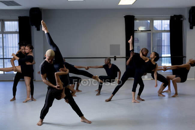 Vue de face d'un mélange de danseurs modernes masculins et féminins en tenue noire pratiquant une routine de danse lors d'un cours de danse dans un studio lumineux, l'homme tient la femme posant à l'envers tandis que d'autres danseurs sont debout en arrière-plan. — Photo de stock