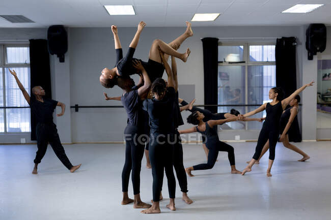 Frontansicht einer multiethnischen Gruppe fitter männlicher und weiblicher moderner Tänzer in schwarzen Outfits, die während eines Tanzkurses in einem hellen Studio eine Tanzroutine praktizieren. Drei männliche Tänzer halten eine Frau über ihren Köpfen. — Stockfoto