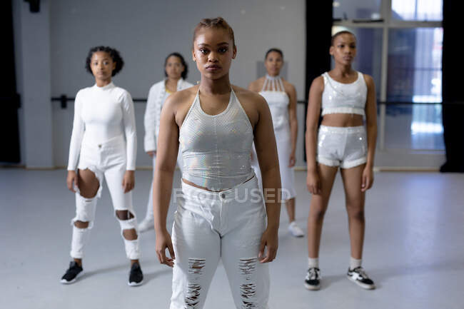 Frontansicht einer multiethnischen Gruppe fitter moderner Tänzerinnen in weißen Outfits, die während eines Tanzkurses in einem hellen Studio eine Tanzroutine praktizieren, auf dem Boden stehend und direkt in eine Kamera schauen. — Stockfoto
