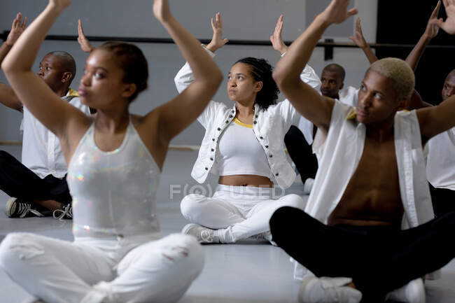 Nahaufnahme einer multiethnischen Gruppe fitter männlicher und weiblicher moderner Tänzer in weißen Outfits, die während eines Tanzkurses in einem hellen Studio eine Tanzroutine praktizieren und mit erhobenen Händen auf dem Boden sitzen. — Stockfoto