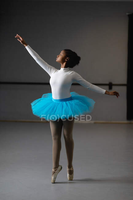 Передній вигляд змішаної раси жіночої балерини в білому трикотажі і синьому турі, танцює в яскравій студії, піднімаючись рукою . — стокове фото