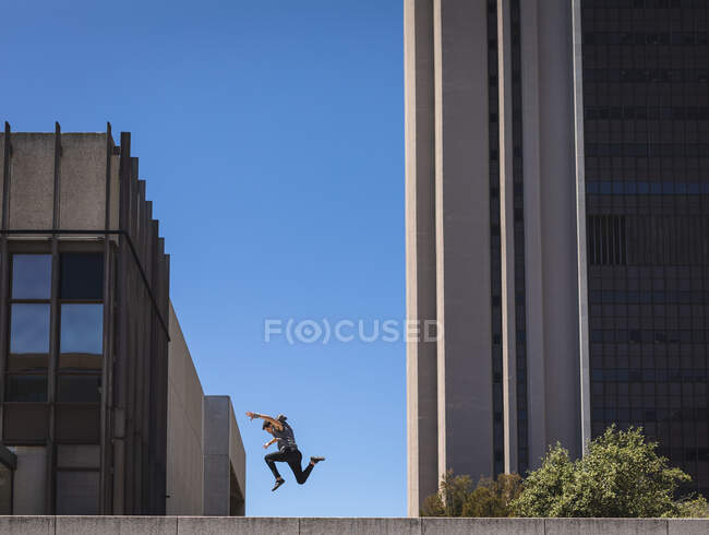 Vista lateral de un hombre caucásico practicando parkour junto al edificio en una ciudad en un día soleado, saltando entre edificios modernos. - foto de stock