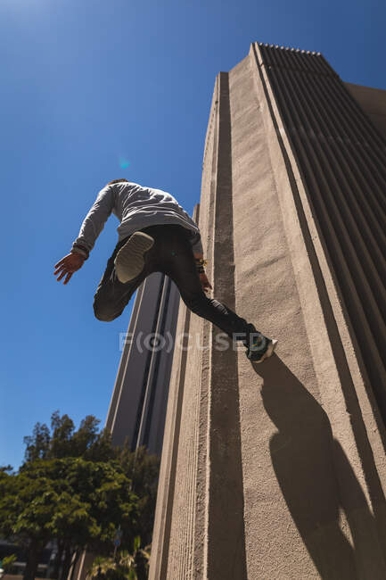 Задній низькокутний вид на кавказького чоловіка, який практикує паркур біля будинку в сонячний день, стрибаючи на бетонну стіну... — Stock Photo