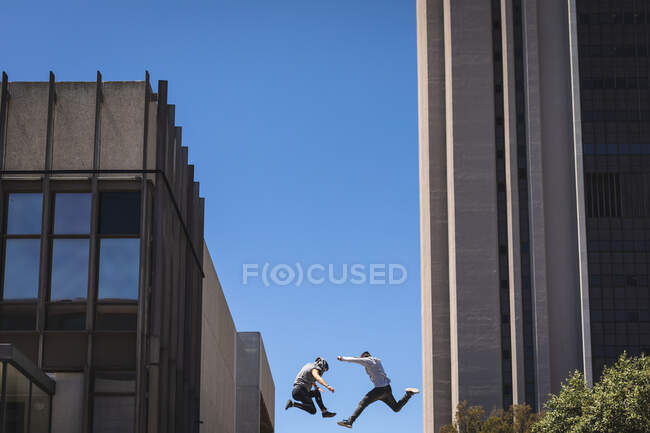 Vue latérale de deux hommes caucasiens pratiquant le parkour près du bâtiment dans une ville par une journée ensoleillée, sautant entre les bâtiments modernes. — Photo de stock