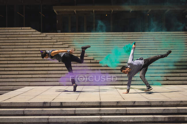 Vista laterale di due uomini caucasici che praticano il parkour vicino all'edificio in una città in una giornata di sole, usando vernici spray sulle scale. — Foto stock