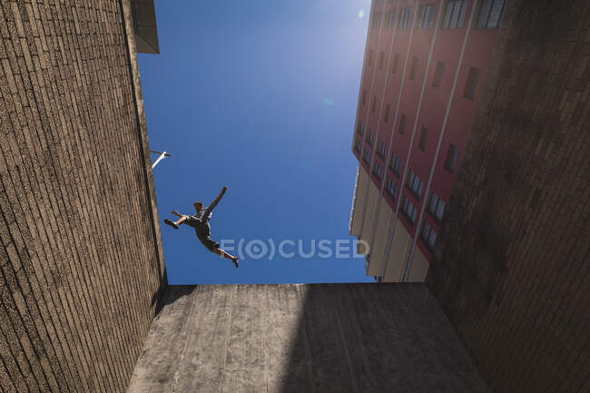 Vue latérale à angle bas d'un homme caucasien pratiquant le parkour près du bâtiment dans une ville par une journée ensoleillée, sautant sur le toit. — Photo de stock