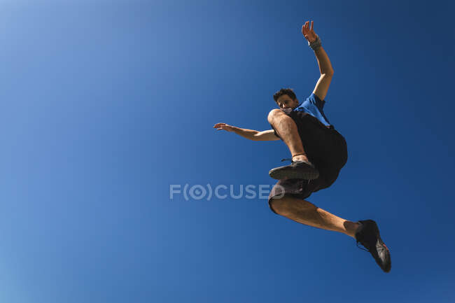 Vista frontal de um homem caucasiano praticando parkour perto do edifício em uma cidade em um dia ensolarado, pulando e espalhando armas. — Fotografia de Stock