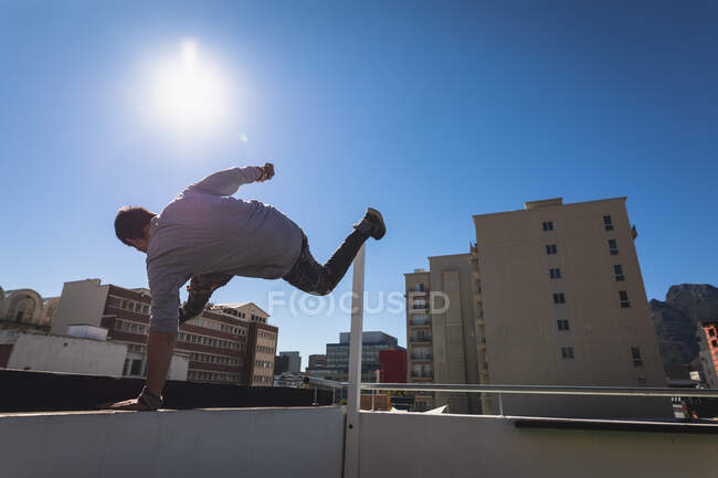 Vue arrière d'un homme caucasien pratiquant le parkour près du bâtiment dans une ville par une journée ensoleillée, sautant sur un toit. — Photo de stock