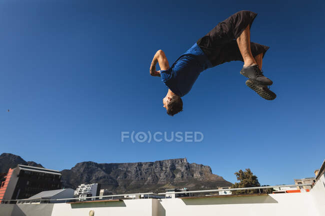 Vista lateral de um homem caucasiano praticando parkour perto do edifício em uma cidade em um dia ensolarado, fazendo um salto mortal em um telhado. — Fotografia de Stock