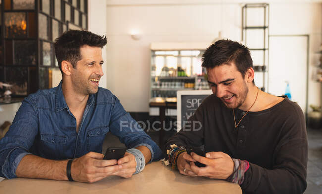Vista frontal de dos hombres caucásicos con ropa casual, sentados junto a una mesa en una cafetería, sonriendo y usando sus teléfonos inteligentes. - foto de stock