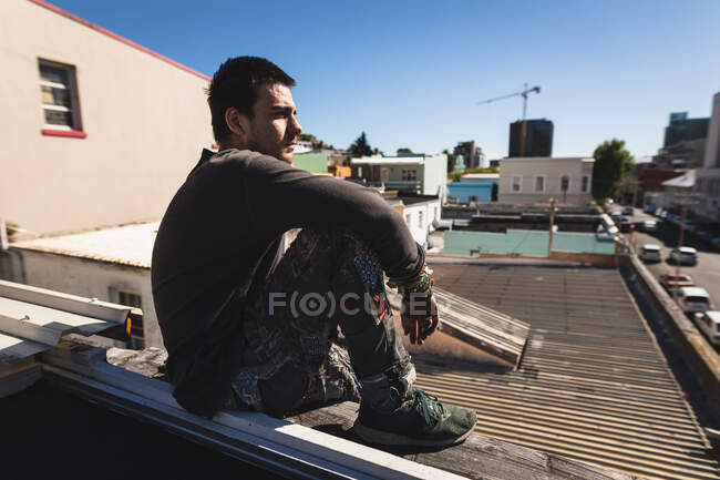 Vista lateral de un hombre caucásico practicando parkour junto al edificio en una ciudad en un día soleado, tomando un descanso, descansando y sentado en una azotea. - foto de stock
