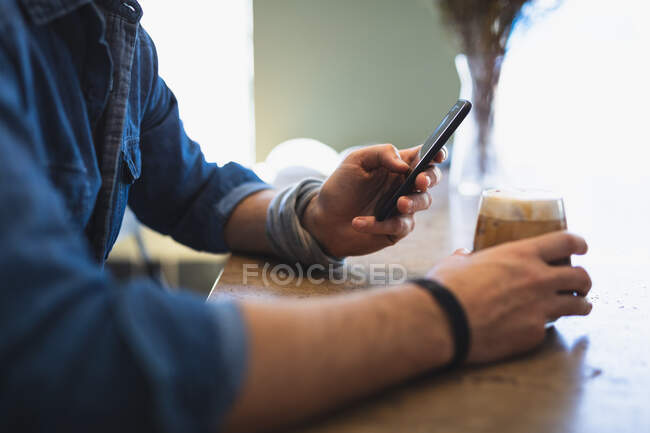 Vista lateral sección media del hombre con ropa casual, sentado junto a una mesa en una cafetería, sosteniendo un vaso de café y usando su teléfono inteligente - foto de stock