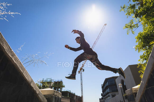 Vue latérale à angle bas d'un homme caucasien pratiquant le parkour près du bâtiment dans une ville par une journée ensoleillée, sautant sur une rampe d'escalier. — Photo de stock