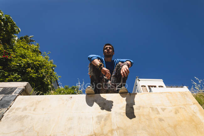Vista frontal de un hombre caucásico practicando parkour junto al edificio en una ciudad en un día soleado, tomando un descanso, descansando y sentado en una pared. - foto de stock