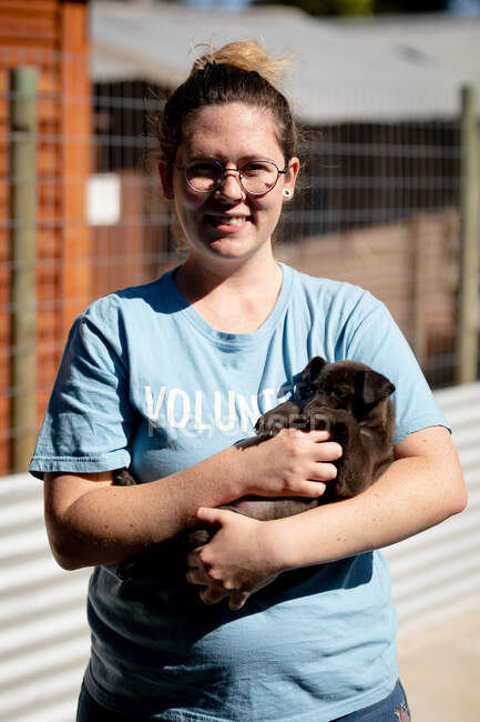 Vista frontal de una voluntaria con gafas y uniforme azul en un refugio de animales sosteniendo a un cachorro rescatado en sus brazos. - foto de stock