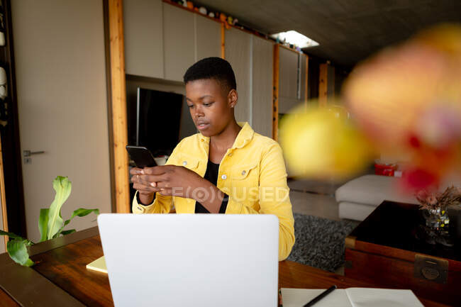 Vista frontal de una mujer afroamericana sentada en su sala de estar en un día soleado, usando una computadora portátil y un teléfono inteligente - foto de stock