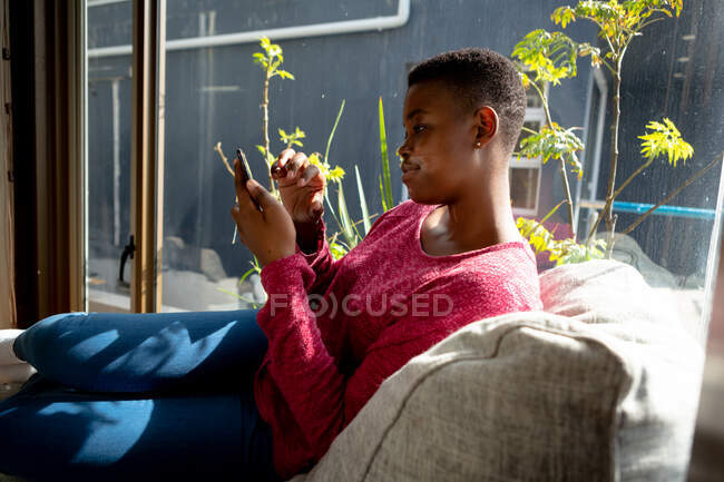 Vista lateral de uma mulher afro-americana sentada em sua sala de estar em frente a uma janela em um dia ensolarado, usando um smartphone — Fotografia de Stock