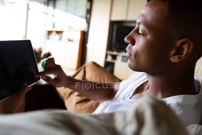 Vista lateral de cerca de un hombre afroamericano pasando el rato en su sala de estar, sentado en un sofá, utilizando una computadora portátil - foto de stock