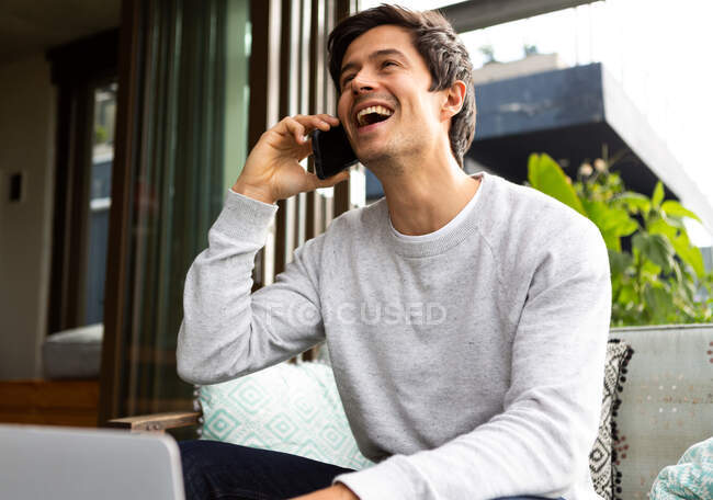 Vista frontale di un uomo caucasico appeso su un balcone in una giornata di sole, seduto su un divano, che parla su uno smartphone e sorride — Foto stock