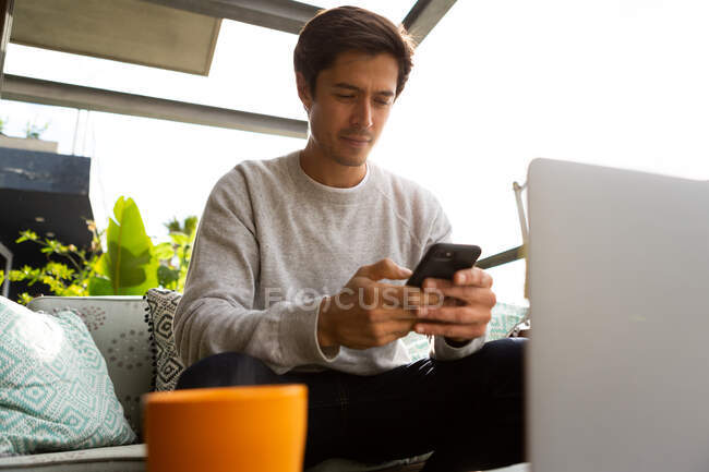 Вид спереди кавказца, болтающегося на балконе в солнечный день, сидящего на диване, используя смартфон — стоковое фото