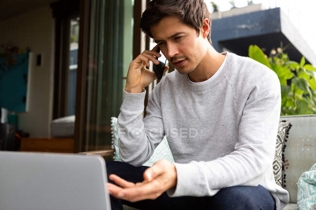 Front view of a Caucasian man hanging out on a balcony on a sunny day, sitting on a sofa, talking on a smartphone — Stock Photo
