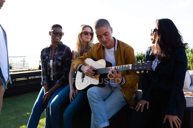Vista frontale di un gruppo multietnico di amici appesi su una terrazza sul tetto in una giornata di sole, sorridente, uno di loro che suona la chitarra — Foto stock
