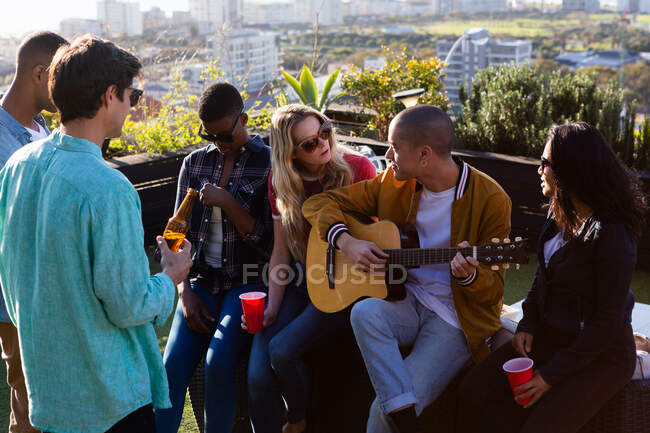 Вид спереду на багатоетнічну групу друзів, які висять на терасі на даху в сонячний день, говорячи, один з них грає на гітарі — стокове фото
